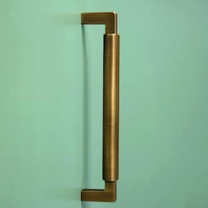 Puede incluir: Un tirador de armario de color bronce sobre un fondo verde azulado claro. El tirador tiene una sección central cilíndrica con extremos cuadrados, creando un diseño moderno y minimalista. El metal tiene un acabado texturizado.