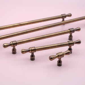 以下が含まれることがあります： シンプルなエレガントなデザインのアンティークブラス製のキャビネットプルが4つあります。プルはサイズ順に配置されており、一番大きいプルが上、一番小さいプルが下にあります。