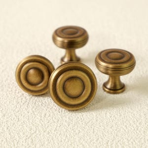 May include: Four antique brass cabinet knobs with a circular design. The knobs have a raised center and concentric rings, with a short, rounded stem. The finish is a warm, aged brass color, suggesting a vintage or classic style.