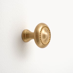 Op de afbeelding: Een messing kastknop met een ronde, met kralen versierde voorkant en een korte, cilindrische steel. De knop is gemonteerd op een wit oppervlak, wat de warme, metalen afwerking en het decoratieve ontwerp benadrukt.