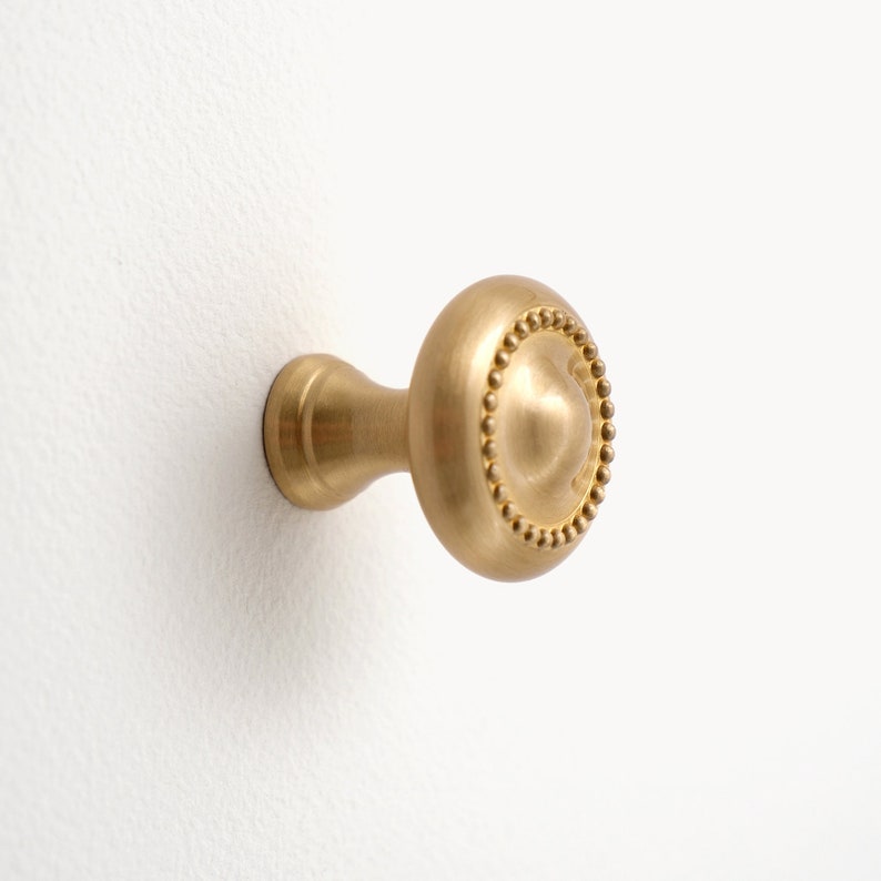 Peut inclure: Un bouton de meuble en laiton avec une face ronde orn&eacute;e de perles et une courte tige cylindrique. Le bouton est fix&eacute; sur une surface blanche, mettant en valeur sa finition m&eacute;tallique chaleureuse et son design d&eacute;coratif.