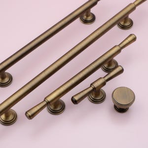 May include: A set of five antique brass cabinet hardware pulls and knobs. The pulls are long and slender with a rounded end, while the knobs are round with a decorative design. The hardware is perfect for adding a touch of vintage style to any home.