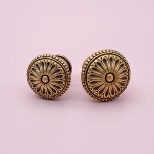 May include: Two gold-toned round cabinet knobs with a detailed floral design. The knobs have a raised centre and a beaded edge.