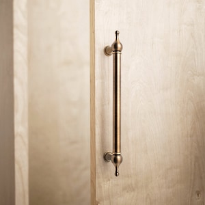 Peut inclure: Une poignée de porte en laiton brossé avec un dessus et un bas arrondis et décoratifs. La poignée est montée sur une porte en bois clair.