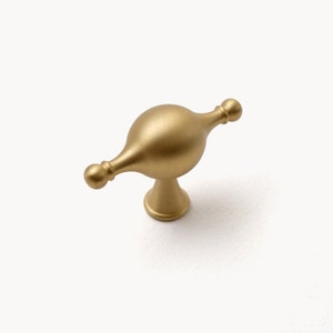 May include: A brushed gold cabinet knob with a rounded, slightly elongated shape and two small knobs on either side.