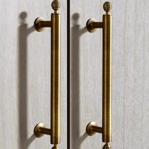 May include: Two gold-toned cabinet door pulls with a decorative knob at the top and a rounded base. The pulls are mounted on a white surface.