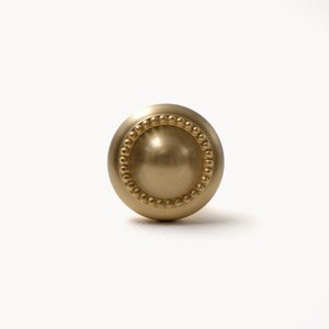 Peut inclure: Un bouton de meuble sph&eacute;rique de couleur dor&eacute;e. Le bouton pr&eacute;sente un cercle central l&eacute;g&egrave;rement encastr&eacute;, entour&eacute; d'un anneau de petites perles en relief. La quincaillerie a une finition lisse et mate, con&ccedil;ue pour les meubles.