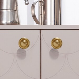 Peut inclure: Deux boutons de meuble dor&eacute;s avec un bord perl&eacute; sont centr&eacute;s sur une porte d'armoire gris clair. Les boutons sont ronds et ont une surface lisse et r&eacute;fl&eacute;chissante. La porte de l'armoire a un motif d&eacute;coratif en forme de coquille.