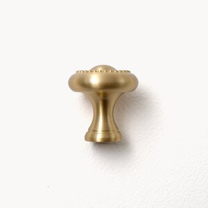 Peut inclure: Un bouton de meuble en laiton avec un dessus arrondi et un bord d&eacute;coratif perl&eacute;. Le bouton a une base &eacute;vas&eacute;e et une finition lisse et polie. La quincaillerie est con&ccedil;ue pour les tiroirs et les armoires.