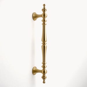 Peut inclure: Une poign&eacute;e de porte dor&eacute;e avec un design orn&eacute;. La poign&eacute;e est fix&eacute;e sur une surface blanche et comporte une longue poign&eacute;e verticale avec des d&eacute;tails d&eacute;coratifs et des fleurons &agrave; chaque extr&eacute;mit&eacute;. La quincaillerie est probablement en laiton ou en m&eacute;tal similaire.