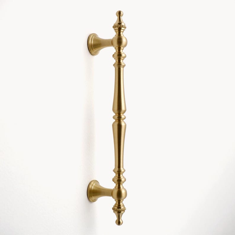 Peut inclure: Une poign&eacute;e de porte dor&eacute;e avec un design orn&eacute;. La poign&eacute;e est fix&eacute;e sur une surface blanche et comporte une longue poign&eacute;e verticale avec des d&eacute;tails d&eacute;coratifs et des fleurons &agrave; chaque extr&eacute;mit&eacute;. La quincaillerie est probablement en laiton ou en m&eacute;tal similaire.