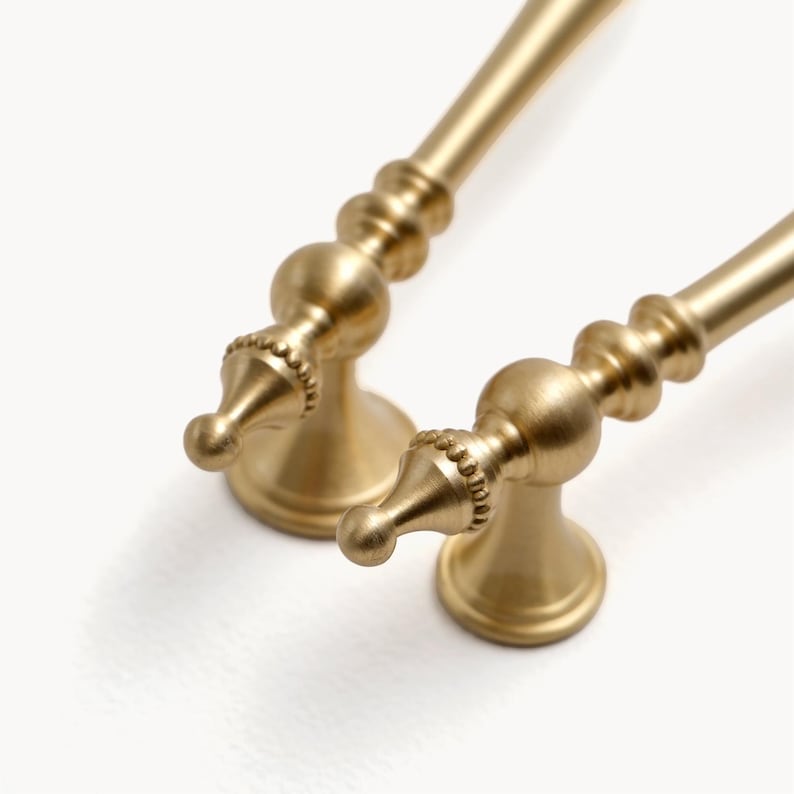 Peut inclure: Deux poign&eacute;es de meuble dor&eacute;es au design classique. Chaque poign&eacute;e pr&eacute;sente une base d&eacute;corative, une section centrale perl&eacute;e et une poign&eacute;e arrondie. Les poign&eacute;es sont en m&eacute;tal et ont une finition mate.