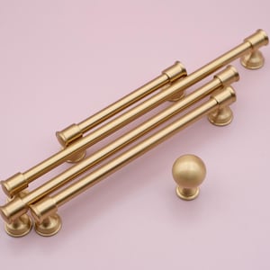 Puede incluir: Cuatro tiradores de armario de color dorado con forma cilíndrica y extremos redondeados. Los tiradores están dispuestos en forma escalonada. También se incluye un solo pomo redondo con una superficie lisa.