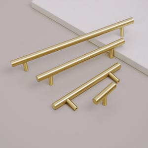 Könnte beinhalten: Eine Sammlung von vier messingfarbenen Schrankgriffen und -knöpfen. Die zylindrischen Griffe haben einen T-förmigen Sockel und sind etwa 5 cm bis 30 cm lang. Die Hardware wird vor einem neutralen Hintergrund präsentiert.