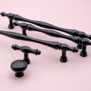 Könnte beinhalten: Ein Satz von fünf schwarzen Schrankknöpfen und -griffen. Die Knöpfe sind rund und die Griffe sind lang und schlank mit einem geschwungenen Design. Die Knöpfe und Griffe sind alle aus Metall.