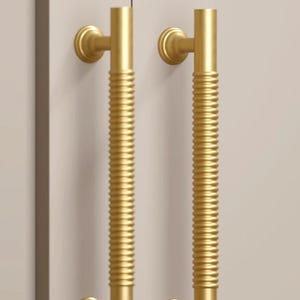 May include: Two gold-colored door handles. Each handle has a cylindrical, ribbed center section and a curved, circular base and top. The handles are mounted on a light-colored surface, suggesting a door or cabinet.