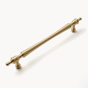 May include: A gold-colored cabinet pull with a straight bar and decorative ends. The hardware is made of metal and has a matte finish. The pull is designed for use on drawers or cabinet doors.