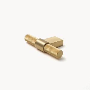 May include: A gold-coloured cabinet pull with a textured, cylindrical handle and a rectangular base. The handle has a knurled pattern for grip. The hardware is designed for furniture and has a modern aesthetic.