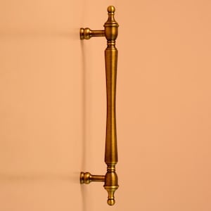 Pode incluir: Puxador de porta de bronze ornamentado com um design vertical e alongado. A pega apresenta detalhes decorativos na parte superior e inferior, com uma pega central em forma de ampulheta. O puxador é montado numa superfície cor de pêssego.