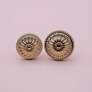 May include: Two gold-toned metal knobs with a floral design. The knobs are round and have a raised centre.