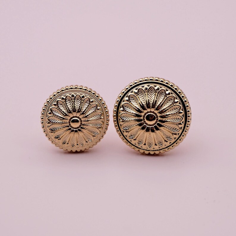 May include: Two gold-toned metal knobs with a floral design. The knobs are round and have a raised centre.