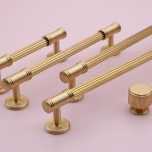 May include: A set of four gold-toned cabinet pulls with a fluted design. The pulls are in various sizes and are arranged in a staggered pattern on a pink background.