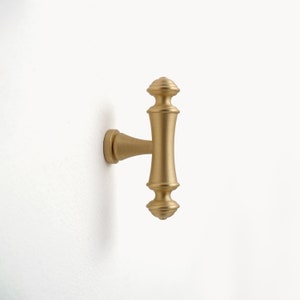 Pode incluir: Uma maçaneta de gabinete em forma de T de cor dourada com um design decorativo. A maçaneta tem uma parte superior e inferior arredondadas e uma barra horizontal reta.