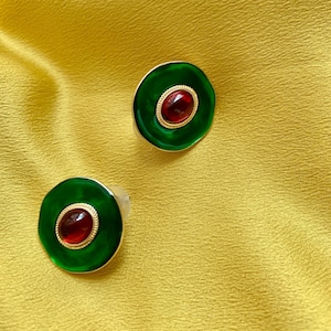 May include: A pair of green and red circular earrings with a gold rim. The earrings have a red gemstone in the center.