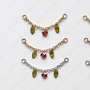 May include: Six delicate chain pendants are displayed. Each pendant has a curved chain with loops at each end. Some have green leaf charms and red flower charms, while others have green leaf charms and clear flower charms. The chains are gold, rose gold, and silver.