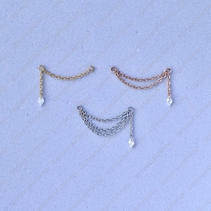 May include: Three pairs of earrings with chain and diamond-shaped charms. One pair is gold, one is rose gold, and one is silver. The earrings are displayed on a light purple surface.