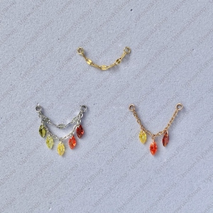 Op de afbeelding: Drie sieradenkettingconnectoren in goud- en zilverkleur. Eén gouden kettingconnector is een gebogen staaf. De andere twee connectoren hebben bungelende, bladvormige edelstenen in geel, oranje en rood.