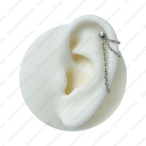 Puede incluir: Un piercing de oreja plateado con un diseño de cadena. La joya está sujeta a un modelo de oreja blanco, destacando los delicados eslabones de la cadena y la estética general. El pendiente está diseñado para la parte superior de la oreja.