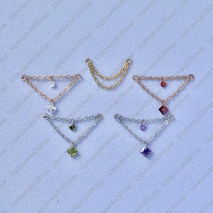 Può includere: Una collezione di gioielli con delicati design a catena. I pezzi includono pendenti a forma di triangolo con gemme pendenti in vari colori, tra cui trasparente, rosso, verde e viola. Alcuni pezzi hanno ciondoli aggiuntivi.