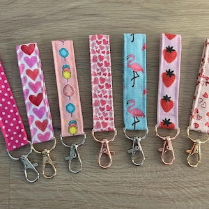 Könnte beinhalten: Sieben bunte Stoffschlüsselanhänger mit verschiedenen Mustern, darunter Herzen, Erdbeeren, Flamingos und Zuckerwatte. Jeder Schlüsselanhänger hat einen silbernen oder goldenen Metallring.