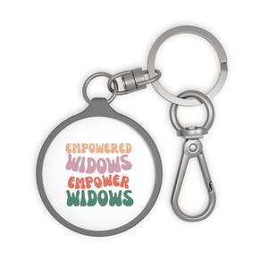 Peut inclure: Un porte-clés argenté avec un charme rond qui dit "Empowered Widows Empower Widows" dans une police rétro avec des couleurs rose, orange et verte.