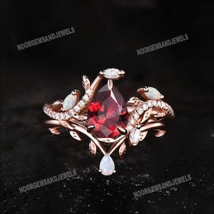 May include: Rose gold ring with a central pear-cut, deep red gemstone, complemented by small white gemstones and opal-like stones. The ring showcases a floral design with leaf and vine detailing.
