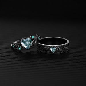 May include: Two rings, one with a heart-shaped, light blue gemstone and smaller teal stones, the other with a heart-shaped stone and leaf design. Both rings are set against a black background.