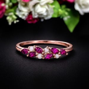 Könnte beinhalten: Roségoldener Ring mit einem Cluster aus fuchsiafarbenen, marquiseförmigen Edelsteinen und kleinen weißen Edelsteinen. Der Ring ist vor einem schwarzen Hintergrund platziert, mit einem verschwommenen Blumenarrangement im Hintergrund.