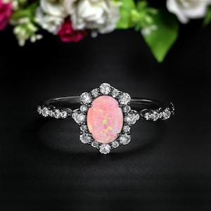 May include: A silver ring with an oval, light pink opal center stone, surrounded by a halo of small, clear gemstones. The ring band is also adorned with small gemstones. The background is black with flowers.