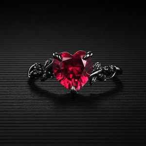May include: A black ring featuring a large, heart-shaped, faceted, red gemstone. The ring has a floral design on the band and around the gemstone. The ring is set against a black background.