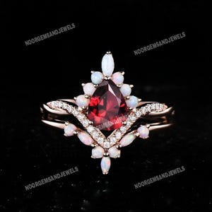 Pode incluir: Um conjunto de anéis em ouro rosa com uma pedra preciosa vermelha escura em forma de pêra como ponto focal, rodeada por pedras brancas menores. O anel tem uma banda delicada e curva com pequenas pedras transparentes.