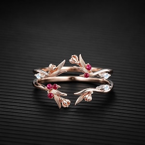 May include: An open-style rose gold ring featuring a botanical design with sculpted leaves and flower buds. The ring is accented with small, round, deep red rubies and clear, round-cut white stones set along the delicate, branch-like metal bands.
