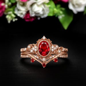 May include: A rose gold ring set featuring a large oval-cut ruby center stone. The ring is accented with small white gemstones and a crown-like design. The set includes a matching band with additional gemstones.
