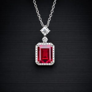 Può includere: Collana d'argento con un pendente rettangolare di pietra rossa, circondato da piccole gemme trasparenti. Sopra il pendente, una gemma quadrata e una rotonda. La catena è sottile e delicata. Un gioiello elegante.