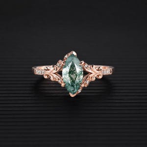 May include: A rose gold ring featuring a marquise-cut, green moss agate gemstone. The ring has a detailed filigree design with small, clear accent stones. The band and setting are a warm, metallic color, set against a dark background.