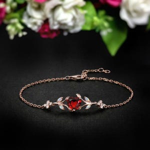 Puede incluir: Una pulsera de oro rosa con una piedra preciosa roja oscura en forma de corazón en el centro. La pulsera está adornada con pequeñas piedras blancas y diseños en forma de hojas a ambos lados del corazón. La cadena es delicada y el cierre es visible.