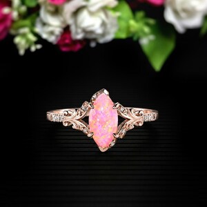 May include: Rose gold ring featuring a marquise-cut pink opal center stone. The ring has a detailed filigree design with small, clear accent stones. The band and setting are a warm, metallic color, set against a dark background.