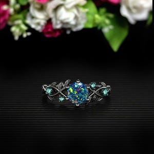 May include: A black ring with a large, multi-colored opal center stone. The ring features a leaf and vine design with small, teal-colored accent stones. The ring is set against a black background with blurred flowers in the background.