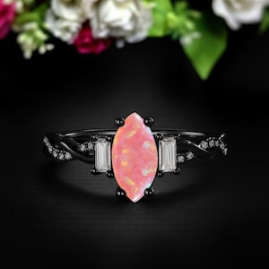 May include: A black ring with a large, pink opal-like center stone. The ring features two rectangular white stones on either side of the center stone. Small white stones are set along the band.