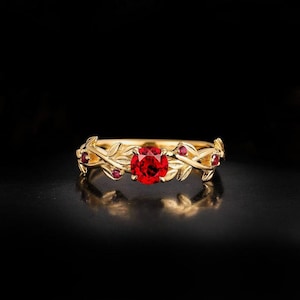 May include: A gold-colored ring with a large, round, red gemstone at the center. The ring features a leaf and vine design with smaller red gemstones. The ring is set against a black background.